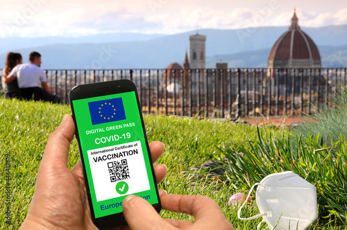 The digital green pass of the European Union with the QR code on the screen of a hand-held mobile phone with a mask and tourists admiring the Cathedral of Florence, Italy. photo