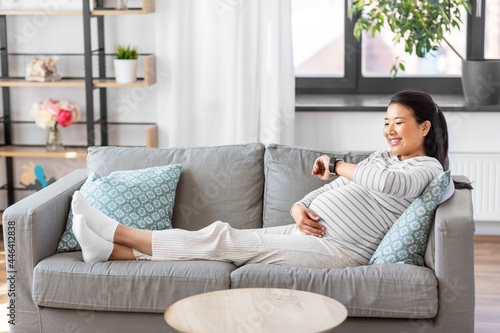 pregnancy, rest, people and expectation concept - happy smiling pregnant asian woman with smart watch sitting on sofa at home