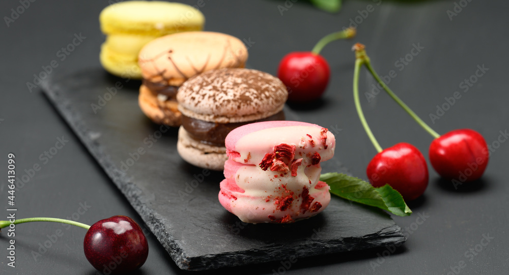 multicolored macarons and ripe red cherries on black wooden background