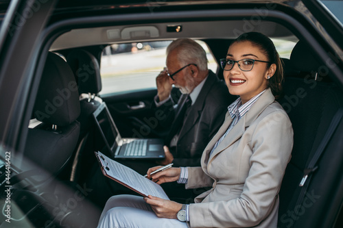 Good looking senior business man and his young woman colleague or coworker sitting on backseat in luxury car. Transportation in corporate business concept.