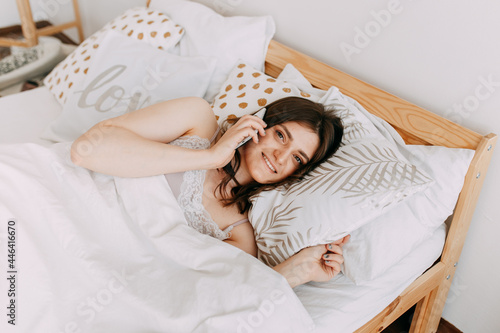 A pretty young freelance woman works communicates takes selfies talks using wireless technologies and a phone lying in bed in a cozy bedroom at home  taken from above  selective focus