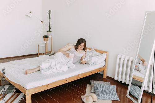 A pretty young freelance woman works communicates takes selfies talks using wireless technologies and a phone lying in bed in a cozy bedroom at home, taken from above, selective focus