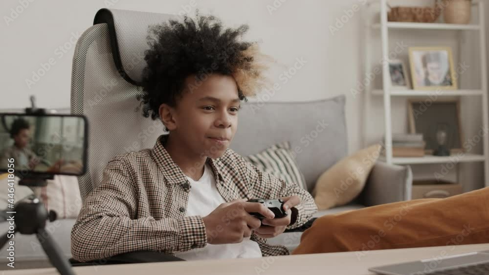 Medium long of cheerful thirteen-year-old boy sitting at desk in room, playing video game using console controller, filming himself on smartphone camera Medium long of cheerful thirteen-year-old boy sitting at desk in room, playing video game using console controller, filming himself on smartphone camera