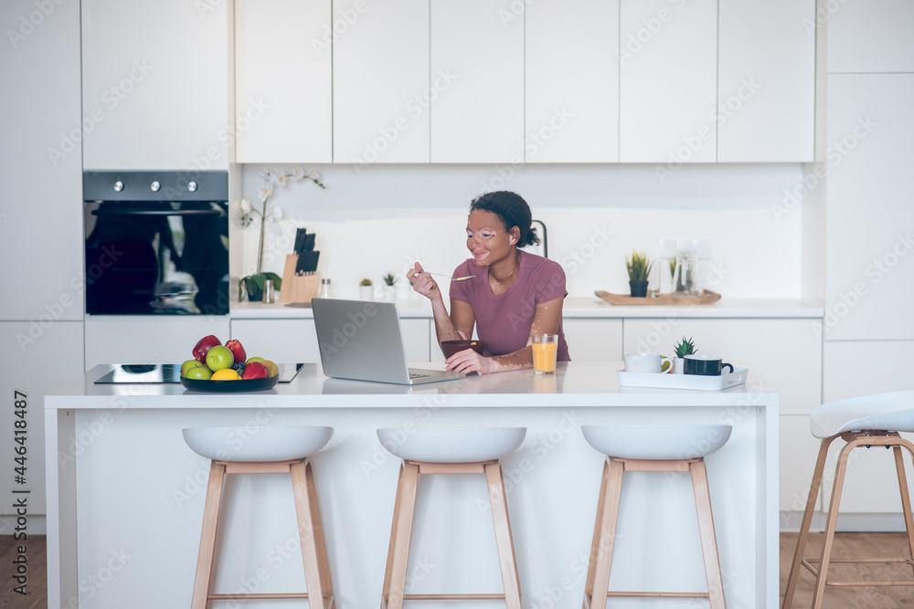 Dark-skinned young woman having breakfast at home and watching somethig on a laptop Dark-skinned young woman having breakfast at home and watching somethig on a laptop