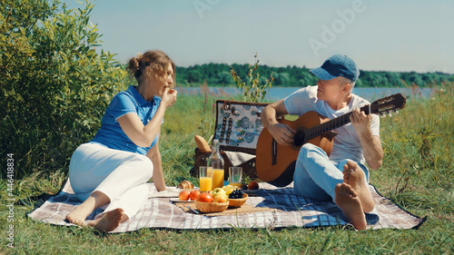 A happy elderly slender couple, a man and a woman, relax with a guitar on a picnic on the river bank on a sunny summer day. The concept of old age, leisure and nutrition.
