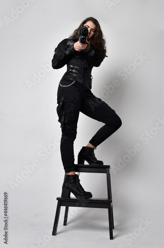 Full length portrait of young woman with natural brown hair, wearing black leather scifi outfit with corset, standing pose on light grey studio background.