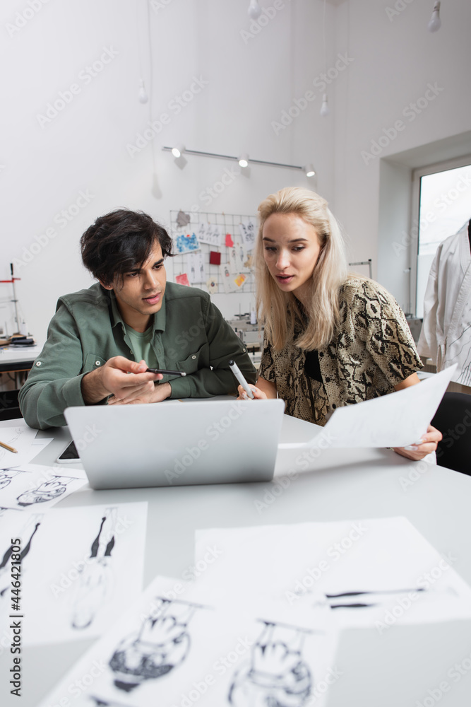 young designer pointing at laptop near blonde colleague and blurred sketches young designer pointing at laptop near blonde colleague and blurred sketches
