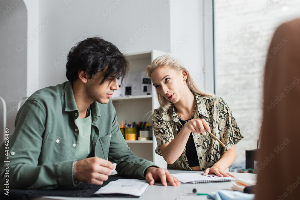 trendy designer pointing with pencil while talking to young tailor in atelier trendy designer pointing with pencil while talking to young tailor in atelier