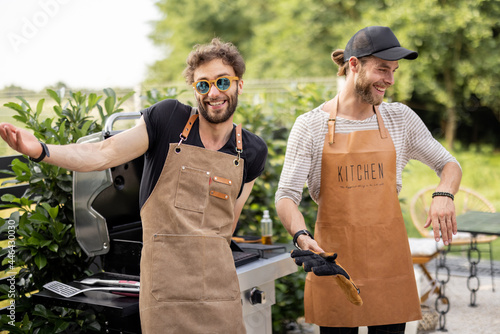 Fotografia, Obraz Portrait of a two handsome male friends in aprons have fun while grilling meat f