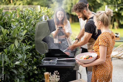 Happy young friends hanging out together, grilling vegetables and meat on a modern grill at picnic. People cooking food outdoors