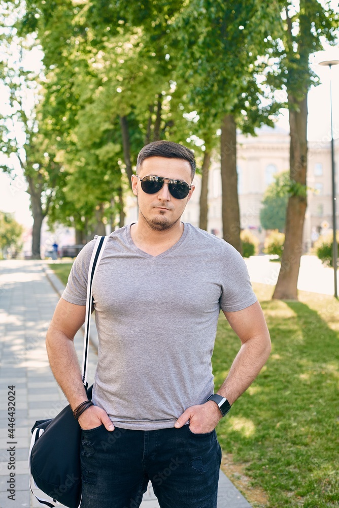 athletic Caucasian man in a gray T-shirt with sunglasses stands against the background of trees athletic Caucasian man in a gray T-shirt with sunglasses stands against the background of trees