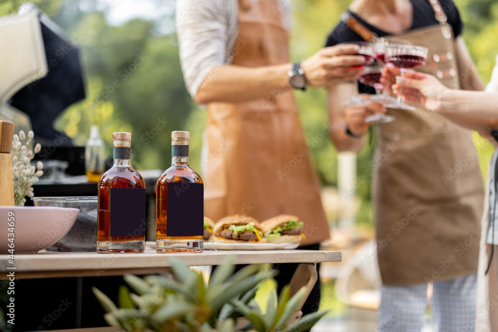 Bottles with liqueur or berry tinctures on a table with burgers, people cheering on a background, close-up on bottles with blank labels for copy paste