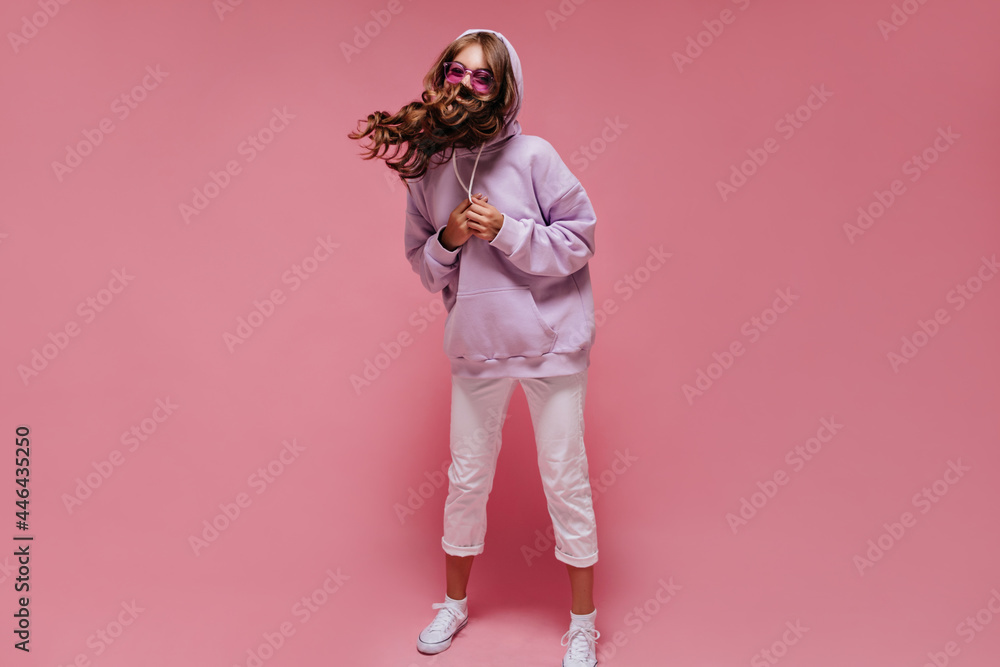 Cheerful woman in purple oversized hoodie and white pants plays hair. Charming girl in sunglasses moves on pink isolated background. Cheerful woman in purple oversized hoodie and white pants plays hair. Charming girl in sunglasses moves on pink isolated background.
