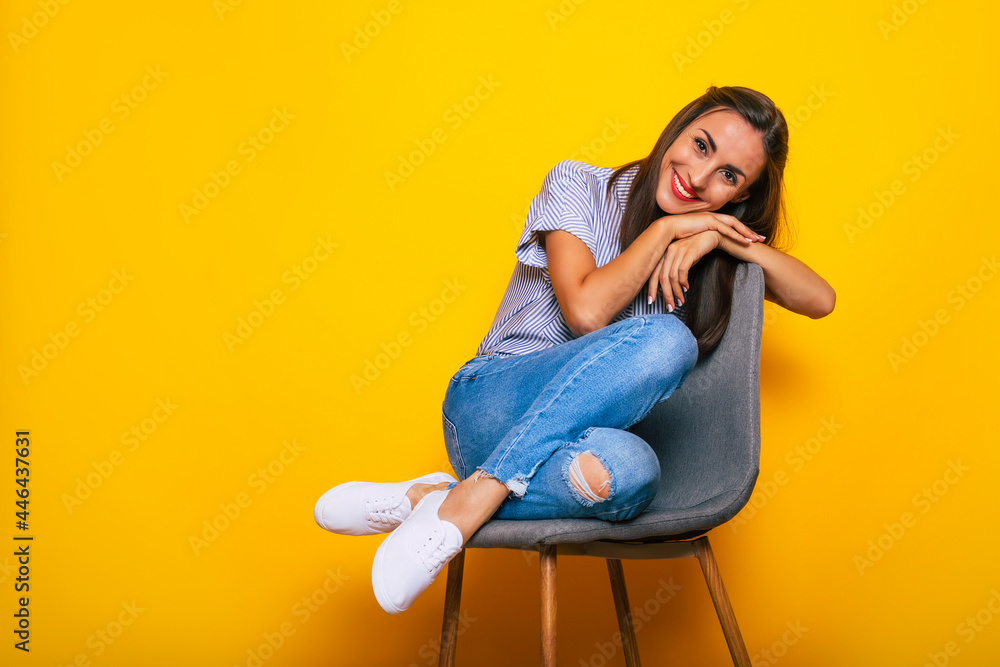 Cute smiling stylish woman is sitting on the chair and posing isolated on yellow background Cute smiling stylish woman is sitting on the chair and posing isolated on yellow background
