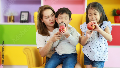 Happy asian family mother  child daughter and son sitting on sofa in colorful modern living room with smiling face while playing magnifying glasses with apples
