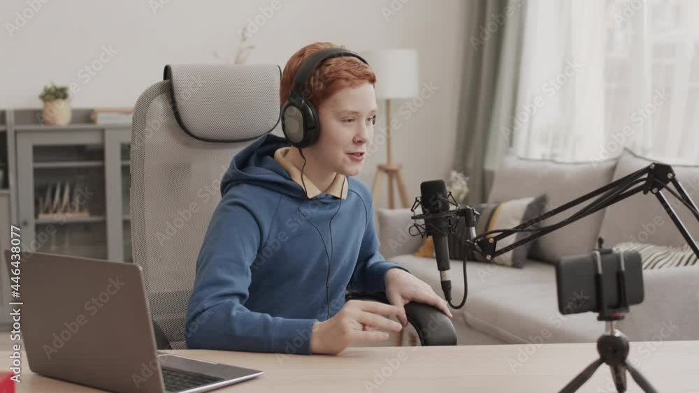 Waist-up of red-haired Caucasian boy wearing blue hoodie and over-ear headphones, sitting at desk in his room, filming video on smartphone for blog Waist-up of red-haired Caucasian boy wearing blue hoodie and over-ear headphones, sitting at desk in his room, filming video on smartphone for blog
