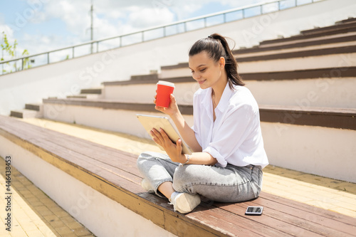 Happy young women with tablet holding coffee paper cup enjoying sunny day sitting in amphitheater.