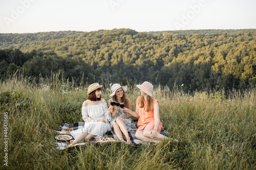 Friendship, picnic and leisure. The company of three gorgeous pretty female friends having fun, cheers and drink wine, and enjoy picnic outdoors. Beautiful hills landscape on the background