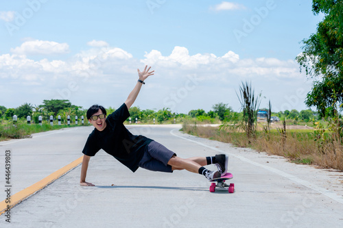 A young Asian man is wearing a black shirt and pants. play skateboard Show the posture of a turn around. On a country road on a sunny day with sky. Looking at the camera, Play surf skate.