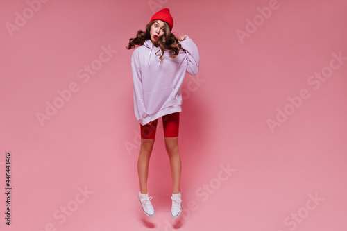 Charming woman in red shorts and oversized purple hoodie jumps on isolated. Brunette girl in hat whistles on pink background.