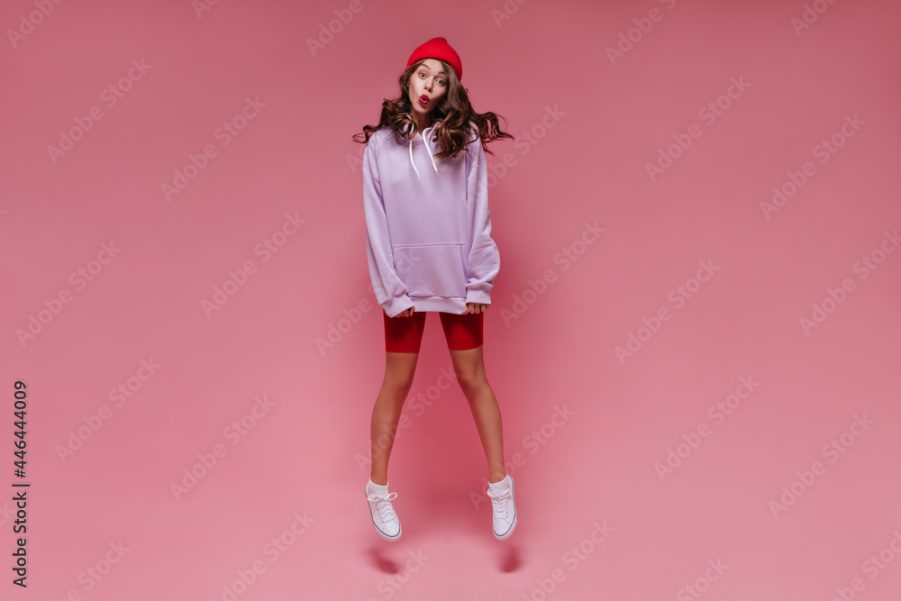 Tanned young girl whistles on pink background. Brunette curly woman in purple oversized hoodie and red shorts jumps on isolated. Tanned young girl whistles on pink background. Brunette curly woman in purple oversized hoodie and red shorts jumps on isolated.