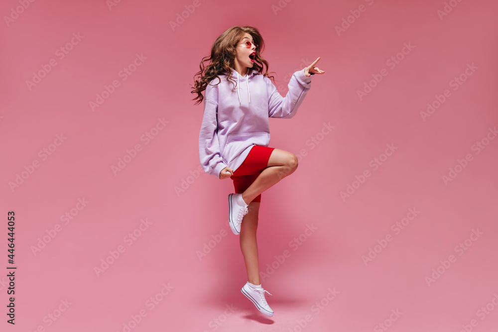 Emotional woman in red cycling shorts and purple hoodie jumps on pink background. Pretty girl in sunglasses moves and points to place for text on isolated. Emotional woman in red cycling shorts and purple hoodie jumps on pink background. Pretty girl in sunglasses moves and points to place for text on isolated.