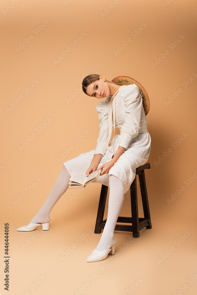 young woman in white dress sitting on stool with book on beige