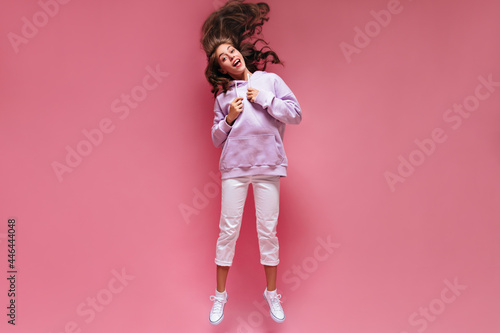 Cool active woman in white pants and purple hoodie jumps on isolated. Full-length portrait of young girl plays hair on pink background.