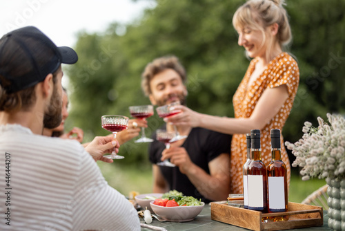 Young friends have a festive lunch outdoors  toasting and drinking wine  spending happy summer time together. Focus on a bottles with blank lables to copy paste on the table