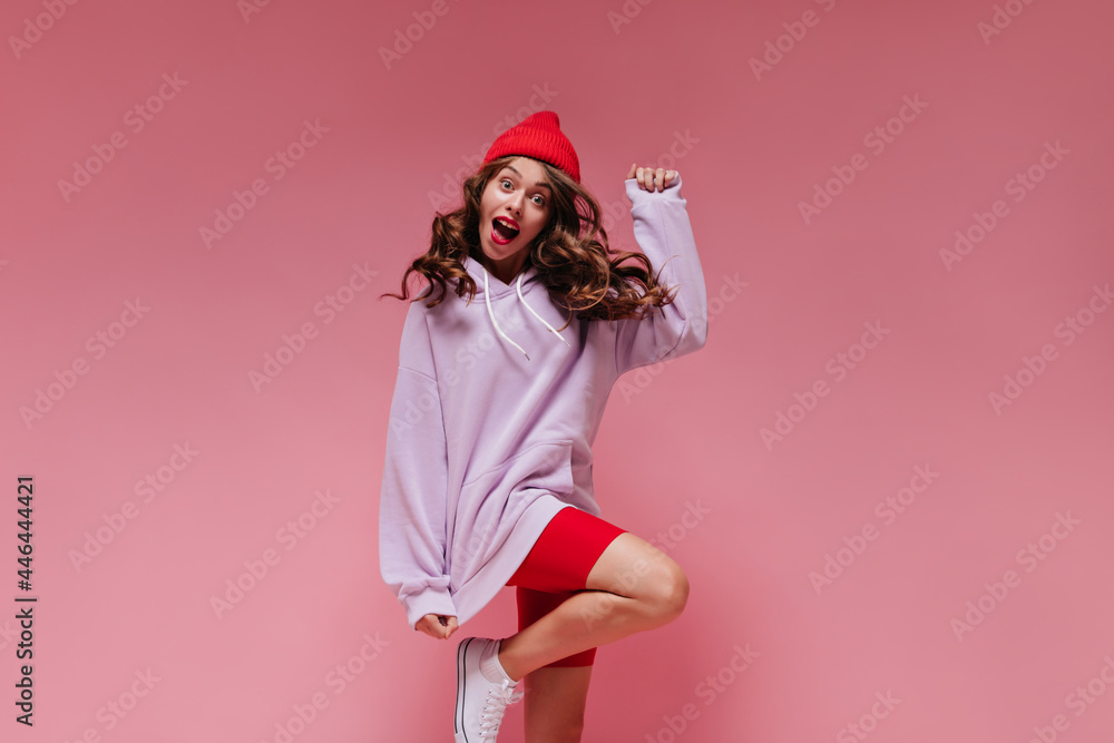 Curly brunette girl in red cycling shorts and purple hoodie jumps on isolated. Cool active woman in hat moves on pink background.