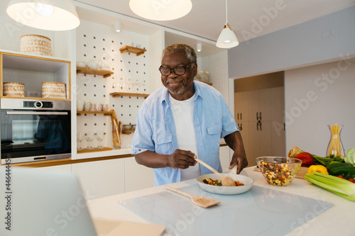Senior African man standing in the kitchen, watching recipe on laptop and preparing a healthy meal.