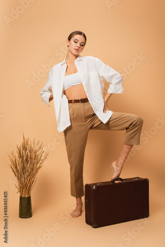 barefoot woman in unbuttoned shirt and pants stepping on vintage suitcase near spikelets on beige background