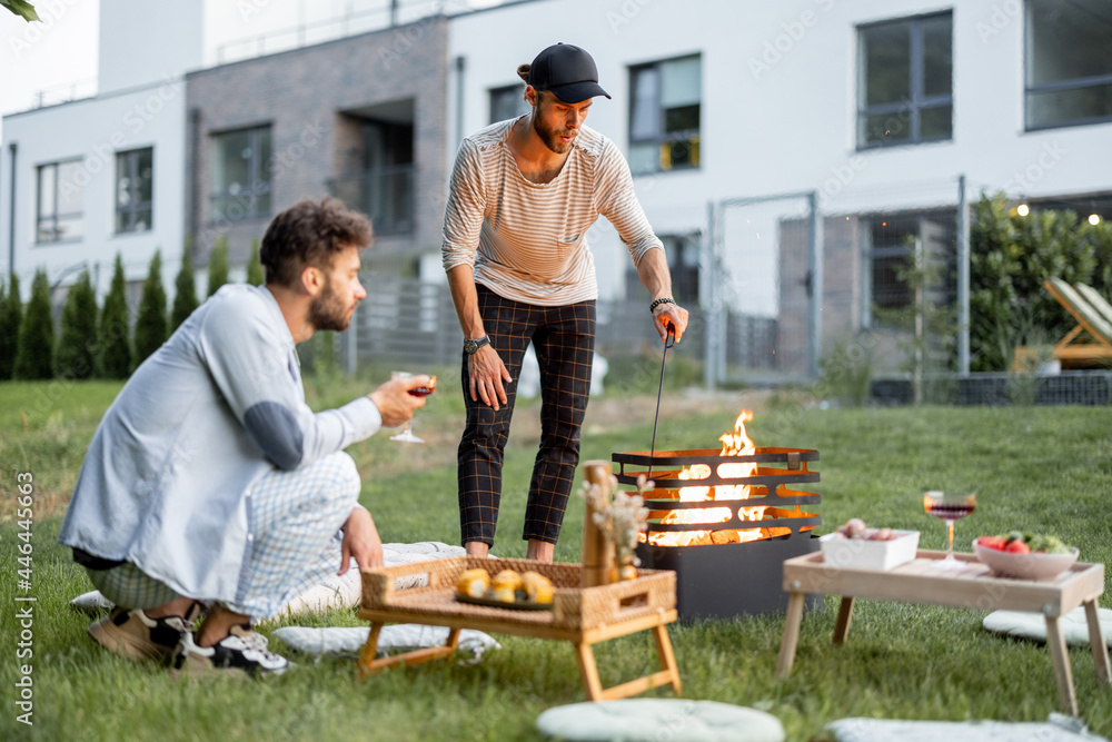 Two male friends have a picnic, making fire at barbeque on the green lawn at backyard of the country houses on the evening