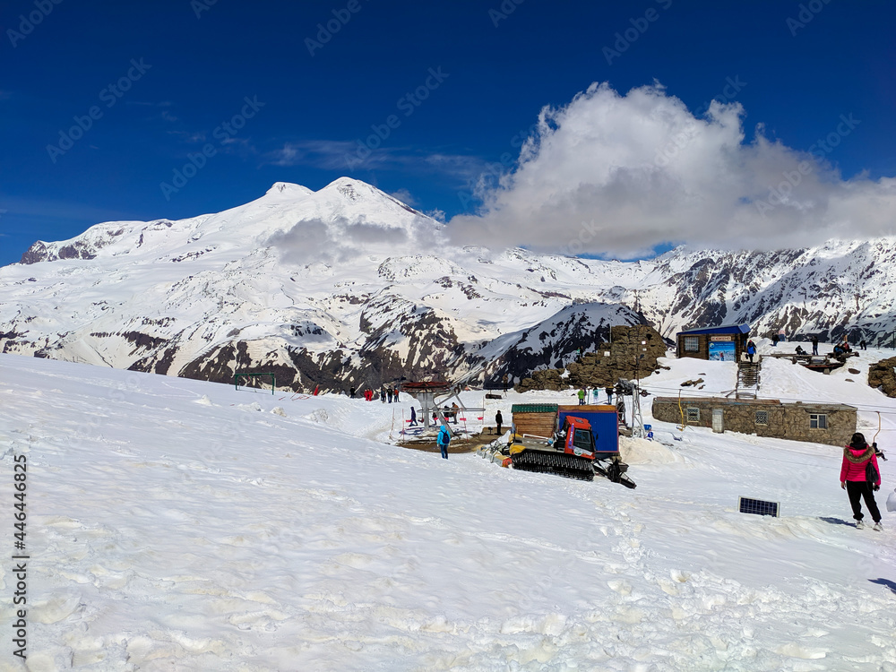 Fototapeta premium Mount Elbrus, Russia - May 10, 2021: view of Mount Elbrus with snow-capped peaks against the blue sky. People walk on Mount Cheget and take pictures against the background of Elbrus.