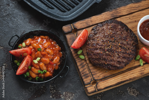 Grilled balkan pljeskavica served with prebranac and red tomatoes, elevated view on a brown stone background, studio shot photo