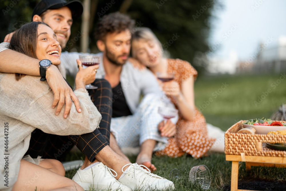 Obraz premium Young friends sit together and hug, talk and drink wine in a close and friendly atmosphere on a picnic in the evening
