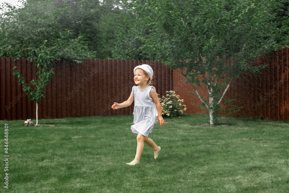 A happy little girl in a dress runs along the garden lawn and plays with a lawn sprinkler.