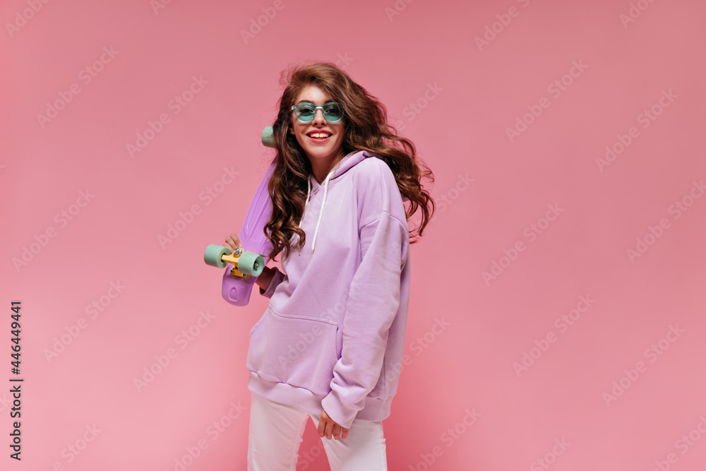 Attractive woman in white pants and purple hoodie looks into camera and smiles. Charming teen girl in green sunglasses holds colorful longboard.