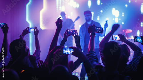 Rock Band with Guitarists and Drummer Performing at a Concert in a Night Club. Front Row Crowd is Record Video on Their Mobile Phones. Party in Front of Bright Colorful Strobing Lights on Stage. 