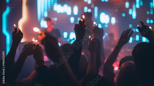 Rock Band Performing a Slow Song at a Concert in a Night Club. Front Row Crowd is Holding Lighters. Silhouettes of Fans Raise Hands in Front of Bright Colorful Strobing Lights on Stage.