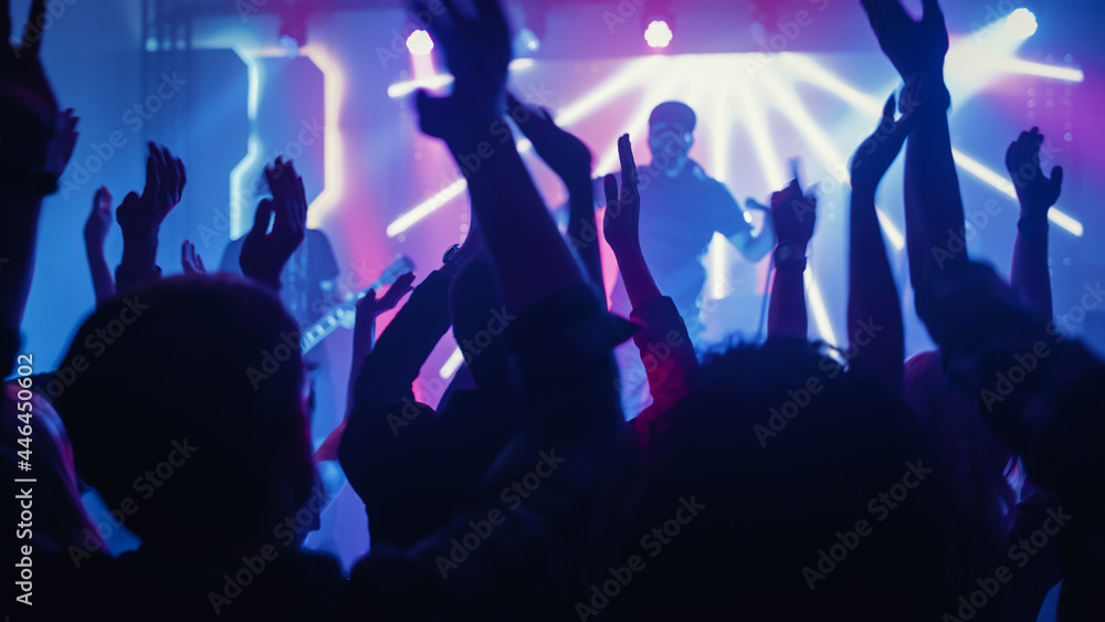 Rock Band with Guitarists and Drummer Performing at a Concert in a Night Club. Front Row Crowd is Partying. Silhouettes of Fans Raise Hands in Front of Bright Colorful Strobing Lights on Stage. 