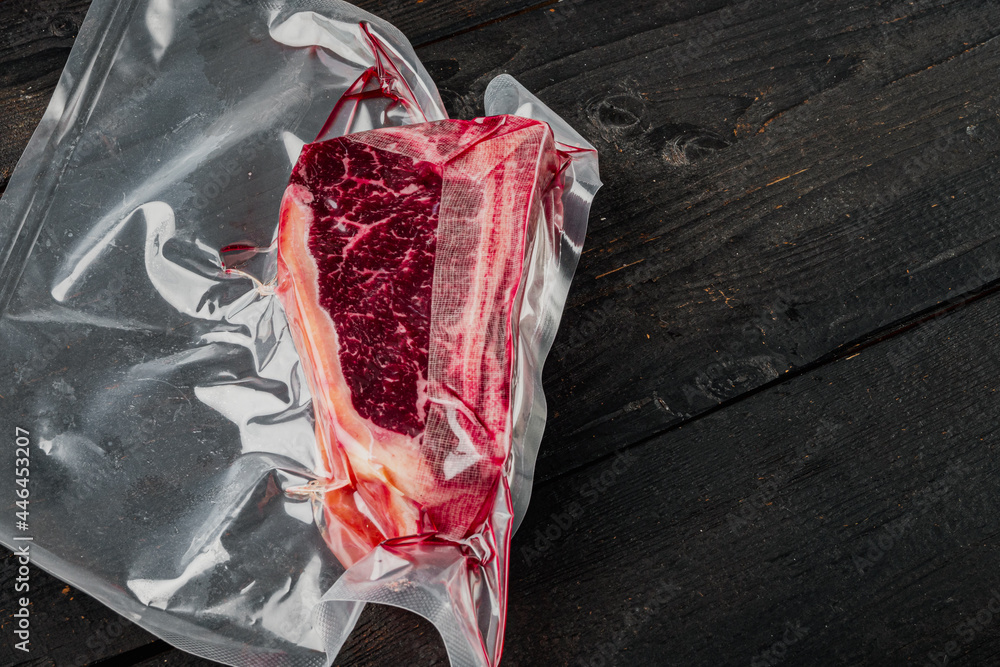 Dry aged beef marbled meat, raw fresh club beefsteak in plastic package, on black wooden table background, with copy space for text