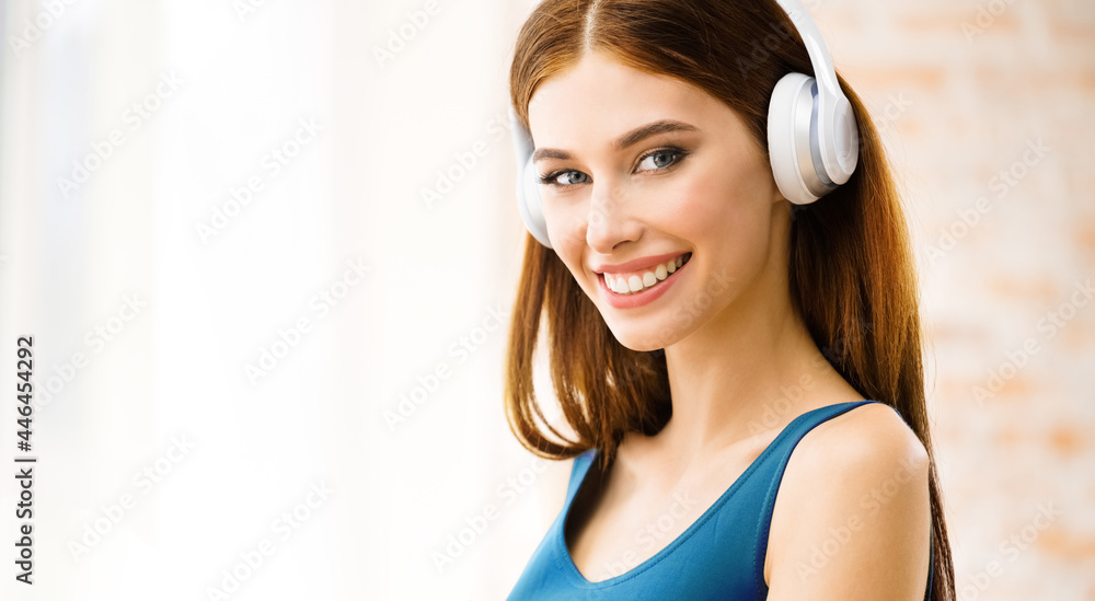 Naklejka premium Portrait image of happy smiling attractive woman listening music in headphones at home. Beautiful girl in casual blue dress standing near window.