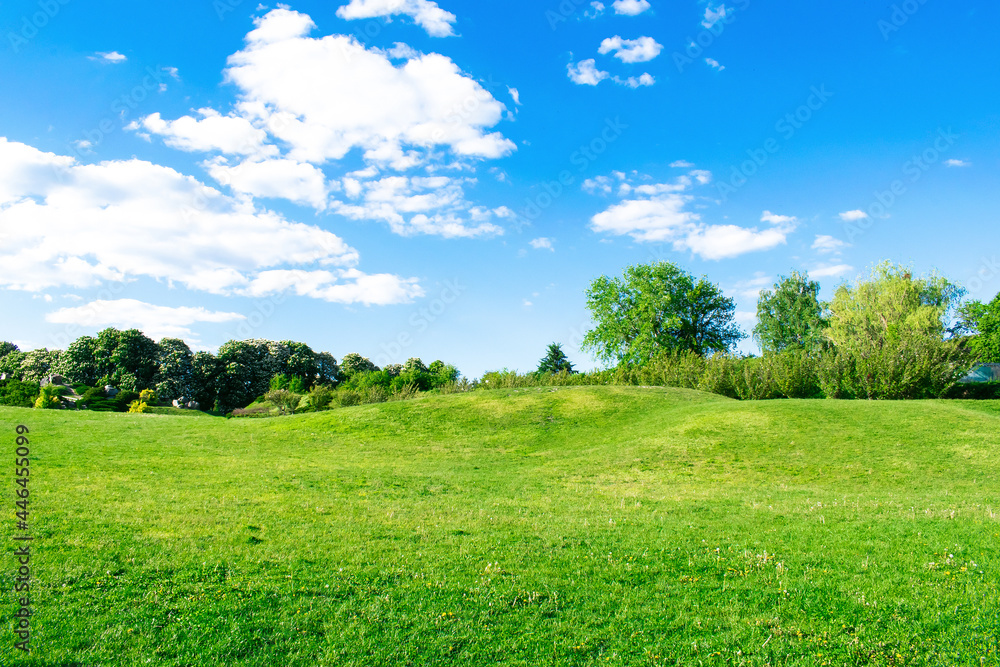 Scenic view of beautiful small mountains, hills, mounds with green grass lawn against bright blue sky with clouds in city park Scenic view of beautiful small mountains, hills, mounds with green grass lawn against bright blue sky with clouds in city park