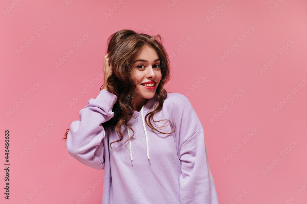 Close-up portrait of brunette curly woman touching her hair. Pretty young girl in purple hoodie smiles and looks into camera on pink background.