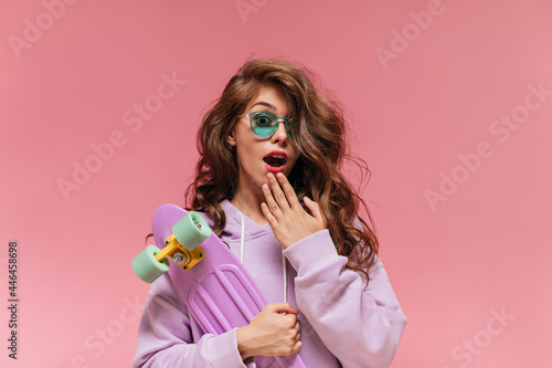 Surprised emotional curly woman holds colorful longboard. Pretty girl in purple hoodie and green sunglasses looks into camera on pink background.