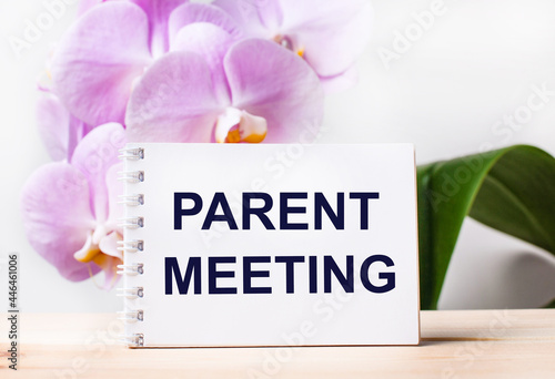 White blank notebook with the text PARENT MEETING on the table against the background of a light pink orchid.