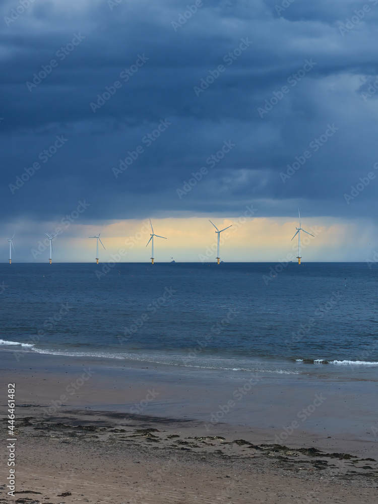The near-shore wind farm at Redcar Beach, ahead of sunlit rent in an ominous, stormy sky, with the rolling waves of the North Sea as foreground. 
