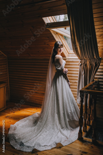 A beautiful bride in a white long dress stands near the window in a wooden room made of logs. Wedding portrait.