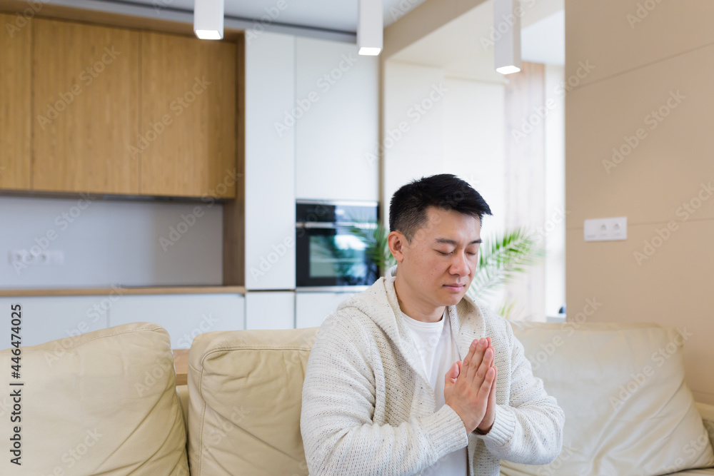 young asian man praying at home alone. Male folding pencil arms and meditating in apartment room. Christian religion traditional spiritual for their hope and believed god forgievness at house